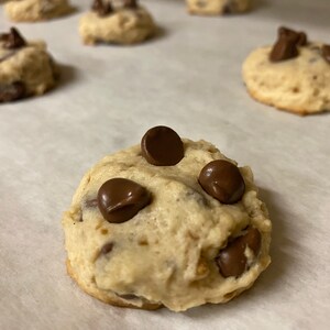 Puede incluir: Primer plano de una galleta de chocolate con chispas con cuatro chispas de chocolate en la parte superior. La galleta es de color marrón claro y tiene una textura ligeramente crujiente. La galleta está sobre papel vegetal blanco.