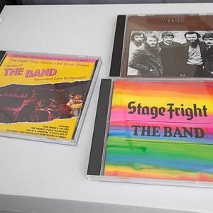May include: Three CD albums by The Band. One album cover is yellow with the text "The Band" and "The Night They Drove Old Dixie Down". Another album cover is brown with a photo of the band. The third album cover is rainbow-coloured with the text "Stage Fright".