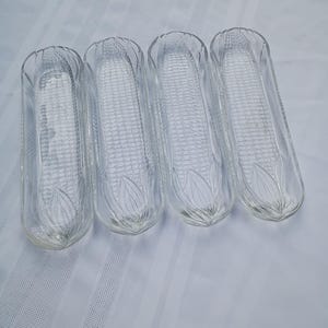 May include: Four clear glass corn-shaped dishes. Each dish has a textured surface resembling corn kernels and a decorative leaf design at the base. The dishes are arranged on a white striped surface.