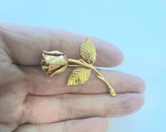 Könnte beinhalten: Eine goldfarbene Rosenbrosche mit detaillierten Blütenblättern und Blättern. Die Brosche ist eine realistische Darstellung einer Rose, mit einem Stiel und Blättern, die sich von der Blüte erstrecken. Die Brosche ist wahrscheinlich aus Metall.