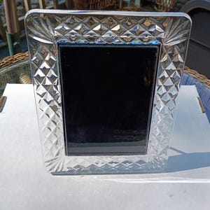 May include: A rectangular picture frame made of clear glass with a faceted diamond-cut pattern along the border. The frame features a solid black rectangular center panel and a reflective, textured surface that catches light across its edges.