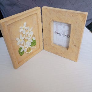 Könnte beinhalten: Ein hellfarbiger, aufklappbarer Fotorahmen mit strukturierter Oberfläche. Eine Seite zeigt ein erhabenes Blumendesign mit weißen Blüten und grünen Blättern. Die andere Seite hat eine quadratische Öffnung mit dem Text "frame-ology".