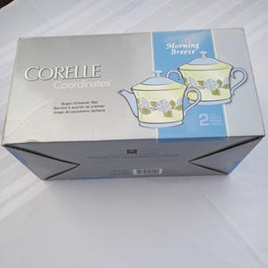 Puede incluir: Una caja de juego de azucarera y cremera Corelle Coordinates. La caja es gris con el logotipo de Corelle Coordinates e información del producto. La caja presenta un gráfico de dos tazas de té con un diseño floral y las palabras "Morning Breeze".
