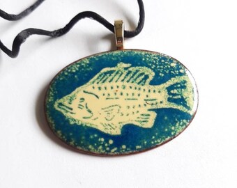 May include: An oval pendant featuring a cream-colored fish graphic set against a mottled blue background. The pendant has a metallic copper-toned edge and is suspended from a thin black cord attached by a gold-toned metal bail.