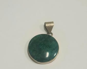 May include: A round pendant with a vibrant green stone set in a silver-colored metal. The stone has a mottled appearance, and the pendant has a simple bail for a chain. The design is simple and elegant.