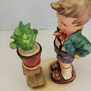 May include: A ceramic figurine of a young person with light brown hair wearing a green jacket, red neckerchief, and purple shorts, standing beside a small green cactus in a red pot on a tan pedestal, all mounted on a round off-white base.