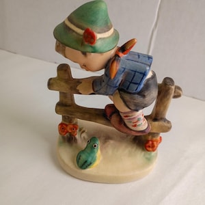 May include: A ceramic figurine depicting a figure wearing a green hat, blue plaid shirt, and dark shorts, climbing over a brown wooden fence. A small green frog sits on the base, which is decorated with small orange flowers on the fence posts.
