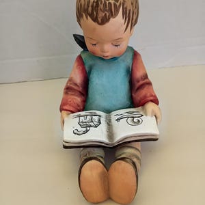 May include: A ceramic figurine of a child with brown hair, wearing a blue vest over a red long-sleeved shirt, sitting and holding an open book. The book pages feature black line drawings of a house with smoke and a snail. The child wears tan shoes.