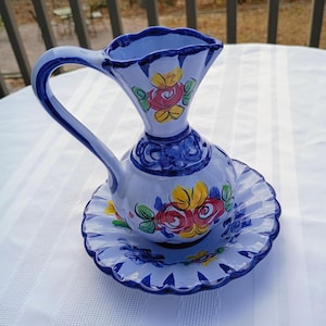 May include: A hand-painted ceramic pitcher and matching saucer set. The pitcher has a curved handle and a flared spout, decorated with blue and floral designs in red, yellow, and green. The saucer has a scalloped edge and a matching floral pattern.