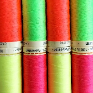 May include: Close-up of vibrant spools of thread in neon shades of red, green, pink, and yellow. The thread is tightly wound on cylindrical spools with white labels. The labels read "100% Polyester".