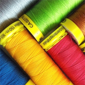 May include: Close-up of spools of embroidery floss in various colours, including yellow, red, green, and blue. The spools have labels with the text "CA 02176", "Interphthal", and "150m - 100%"