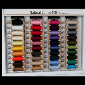 May include: A wall display of 100 meter spools of natural cotton thread in various colors. The spools are arranged in rows and columns, with each spool labeled with a number. The label "Natural Cotton 100 m" is at the top of the display.