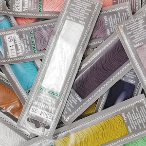 K&ouml;nnte beinhalten: Sortiment an bunten Stickgarn-Spulen in verschiedenen Farbt&ouml;nen von Blau, Gr&uuml;n, Gelb, Orange, Rosa, Lila und Wei&szlig;. Jede Spule ist mit dem Markennamen "Madeira Moulin&eacute;" und der L&auml;nge des Garns in Yards und Metern gekennzeichnet.