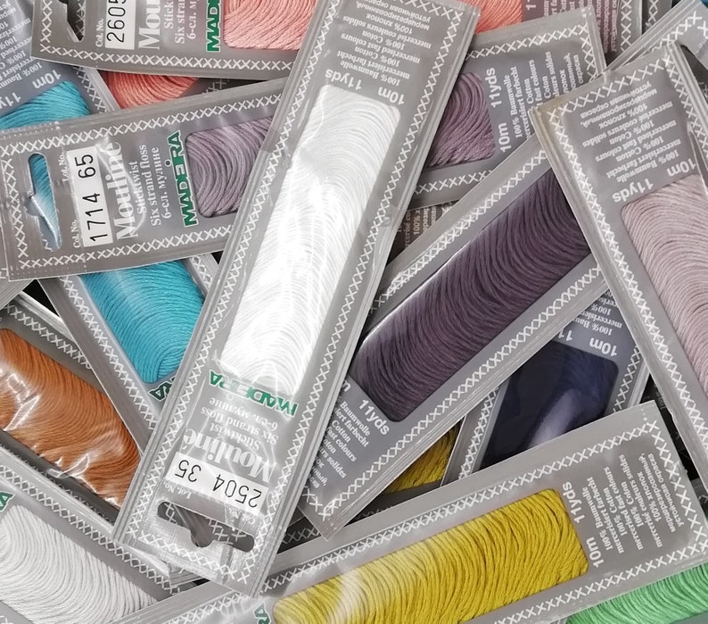 K&ouml;nnte beinhalten: Sortiment an bunten Stickgarn-Spulen in verschiedenen Farbt&ouml;nen von Blau, Gr&uuml;n, Gelb, Orange, Rosa, Lila und Wei&szlig;. Jede Spule ist mit dem Markennamen "Madeira Moulin&eacute;" und der L&auml;nge des Garns in Yards und Metern gekennzeichnet.