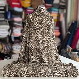 May include: A bolt of fabric with a leopard print design. The fabric is draped over a mannequin and table, showcasing the brown and tan spots on a cream background. The fabric appears soft and flowing, suitable for apparel or home decor.