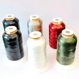 May include: Six spools of Gütermann Outdoor 40 thread in black, silver, red, white, and olive green. Each spool features a white top label with the text Outdoor 40, 400 m, and 437 yds. The thread is wound onto cylindrical plastic spools with white bases.