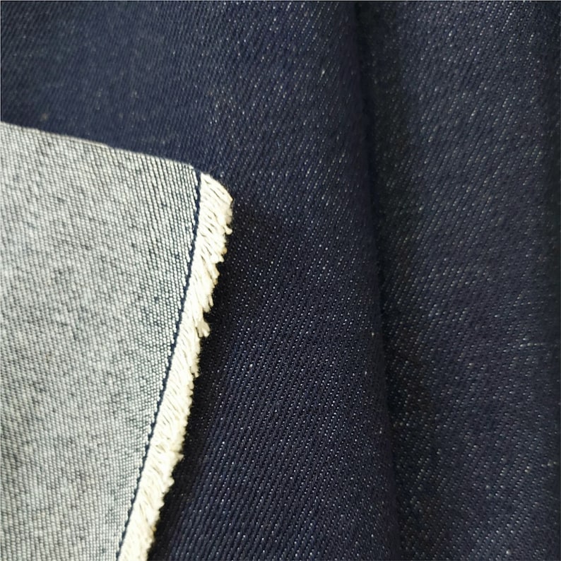 Op de afbeelding: Close-up van donkerblauwe denim stof met een witte selvedge rand. De stof is geweven met een diagonaal keperpatroon.