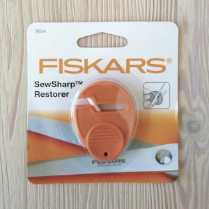 May include: An orange Fiskars SewSharp Restorer tool in its packaging. The package is white and orange with the Fiskars logo and product name. The tool is a small, orange, oval-shaped device with a slot for sharpening scissors.