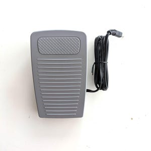 May include: A grey foot pedal with a black cord. The pedal has a textured surface and a rectangular shape.
