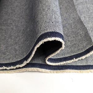 Op de afbeelding: Close-up van een donkerblauwe denim stof met een witte en marineblauwe bies. De stof is dubbelgevouwen en de randen zijn rafelig.
