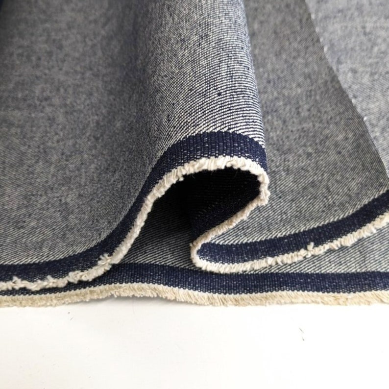 Op de afbeelding: Close-up van een donkerblauwe denim stof met een witte en marineblauwe bies. De stof is dubbelgevouwen en de randen zijn rafelig.