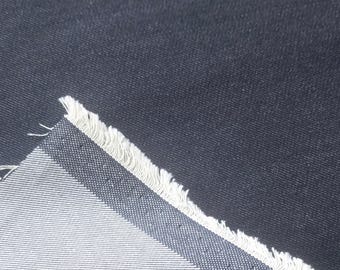 Może przedstawiać: Kawałek ciemnogranatowej tkaniny o widocznej splocie skośnym. Materiał jest złożony, ukazując jaśniejszy, jasnoszary spód z wyraźnym diagonalnym wzorem splotu oraz postrzępioną, surową krawędź z luźnymi białymi nitkami.