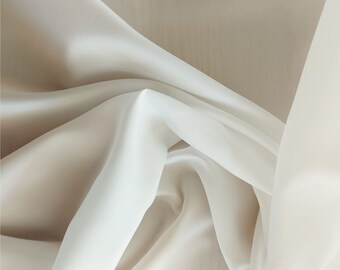 Op de afbeelding: Een close-up van een doorschijnende, witte stof gedrapeerd in zachte plooien. De stof heeft een gladde, zijdeachtige textuur en een delicate, doorschijnende uitstraling.