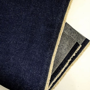 Op de afbeelding: Close-up van een donkerblauwe denim stof met een rauwe rand. De stof is dubbelgevouwen en de rauwe rand is aan beide kanten zichtbaar.