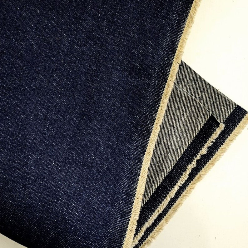Op de afbeelding: Close-up van een donkerblauwe denim stof met een rauwe rand. De stof is dubbelgevouwen en de rauwe rand is aan beide kanten zichtbaar.