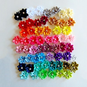 May include: Assortment of 24 handmade satin ribbon flowers with rhinestone centers. The flowers are arranged in rows, with each row featuring a different color. The colors include white, gray, yellow, orange, red, pink, purple, blue, green, and brown.