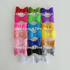 May include: Assortment of 18 sequin bows in various colours, including red, pink, green, blue, yellow, orange, gold, silver, black, and white. The bows are arranged in three rows of six bows each. The text "Once Upon A Time Tutus" is printed below each row of bows.