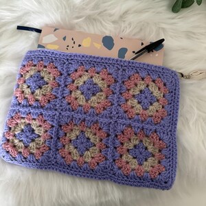 May include: A purple crocheted zippered pouch with a granny square pattern in pink and beige. The pouch is open and a notebook is inside.