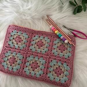 May include: A pink crocheted pouch with a geometric pattern, featuring squares of pink, beige, and blue yarn. The pouch is lying on a white fluffy surface with crochet hooks and scissors nearby.