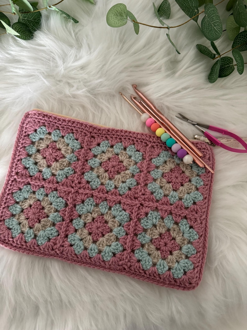 May include: A pink crocheted pouch with a geometric pattern, featuring squares of pink, beige, and blue yarn. The pouch is lying on a white fluffy surface with crochet hooks and scissors nearby.