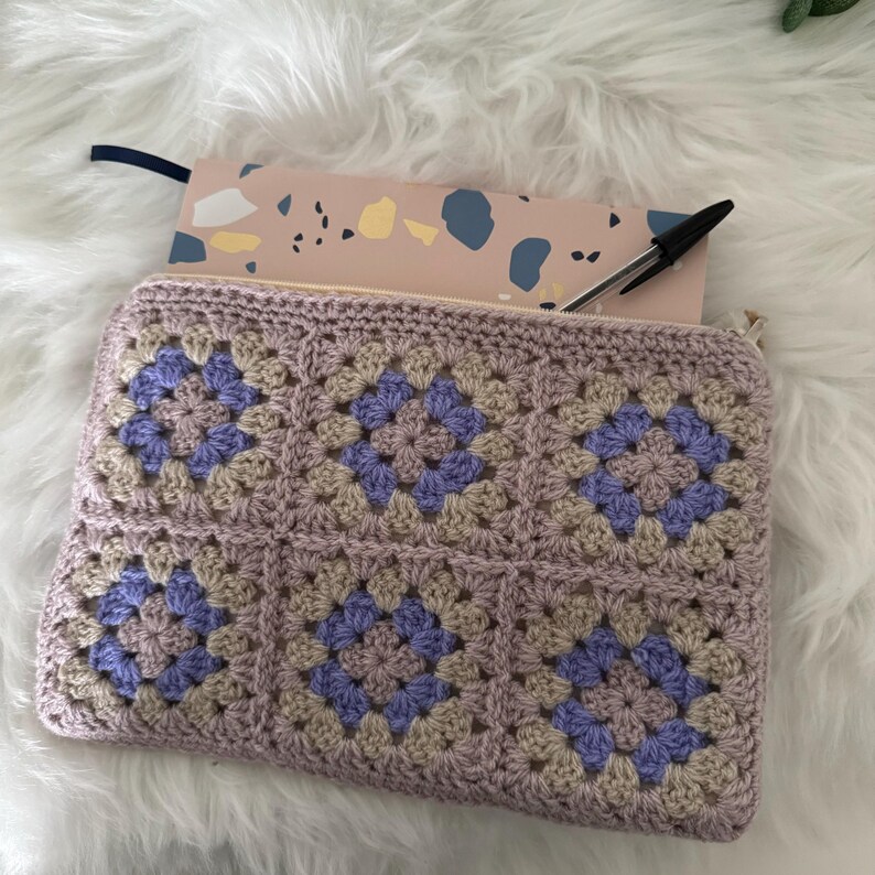 May include: A crocheted pouch with a zipper closure, featuring a granny square pattern in shades of beige and purple. The pouch is lying on a fluffy white surface.