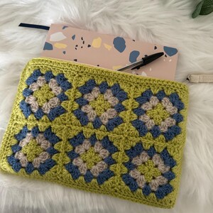 May include: A green, blue and beige crocheted zippered pouch with a granny square pattern. The pouch is open and a notebook is inside.