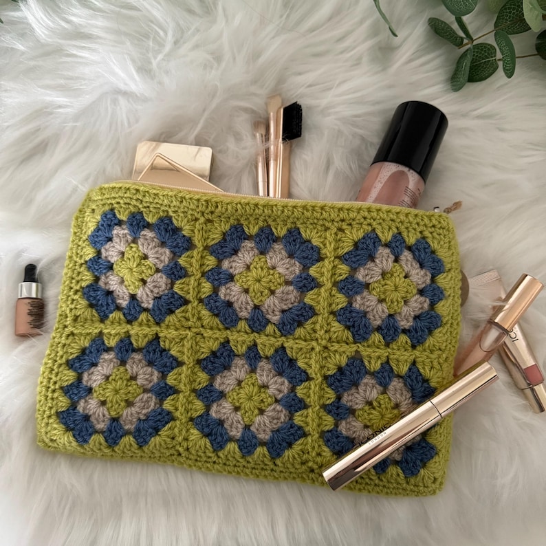 May include: A green, blue and beige crocheted makeup bag with a zip closure. The bag is filled with makeup brushes and other cosmetics.