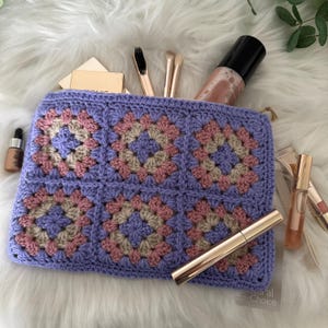 May include: A purple crocheted pouch with a granny square pattern in pink and beige. The pouch is filled with makeup brushes, a bottle of makeup setting spray, and a tube of mascara.