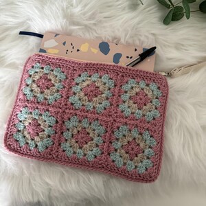 May include: A pink crocheted zippered pouch with a light blue and beige granny square pattern. The pouch is lying on a white fluffy surface with a notebook and pen inside.