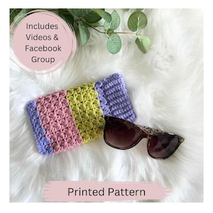 May include: A crocheted glasses case with stripes of pink, green, and purple. The case is lying on a white fluffy surface with a pair of sunglasses next to it. The text "Includes Videos & Facebook Group" and "Printed Pattern" are visible in the image.