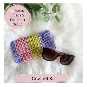 May include: Crochet kit for a glasses case with a purple, pink and green striped design. The kit includes videos and a Facebook group. The case is lying on a white fluffy surface with a pair of sunglasses next to it.