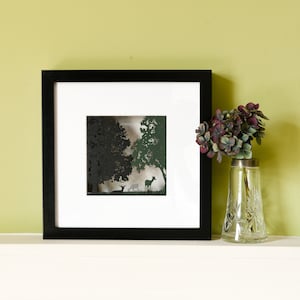 May include: A black framed picture with a white mount and a paper cut-out design of a woodland scene with deer. The woodland is depicted in shades of green and black. The picture is on a white shelf with a vase of purple flowers.