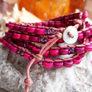 May include: A multi-strand wrap bracelet with pink wooden beads and a brown leather cord. The bracelet has a white button closure.
