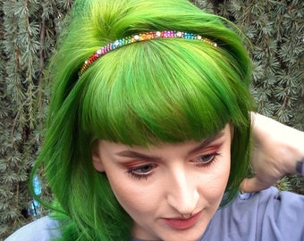 May include: A thin headband featuring a row of small, multi-colored rhinestones in a rainbow gradient pattern. The headband is worn across the top of bright green hair, with the sparkling crystals catching the light against the vibrant hair color.