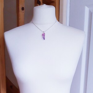May include: A silver necklace with a purple and pink crystal pendant. The crystal is an elongated, irregular shape, set in a silver bezel. The necklace is displayed on a white mannequin, showcasing the jewellery.