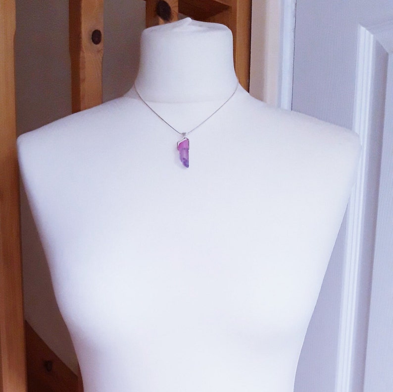 May include: A silver necklace with a purple and pink crystal pendant. The crystal is an elongated, irregular shape, set in a silver bezel. The necklace is displayed on a white mannequin, showcasing the jewellery.