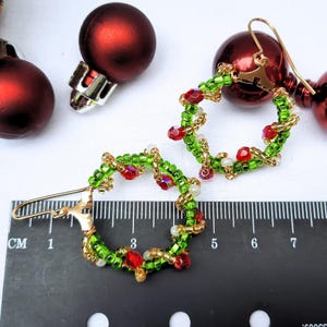May include: Handmade beaded Christmas earrings. The earrings are green, red, and gold, with a hoop design. The earrings are approximately 3.8 cm in diameter. Red Christmas baubles are in the background.