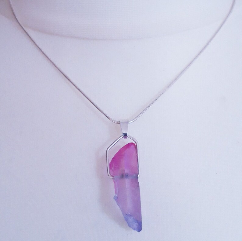 May include: A silver necklace with a pendant featuring a pink and purple crystal. The crystal is a long, irregular shape, with a silver metal frame. The chain is a thin, silver snake chain.