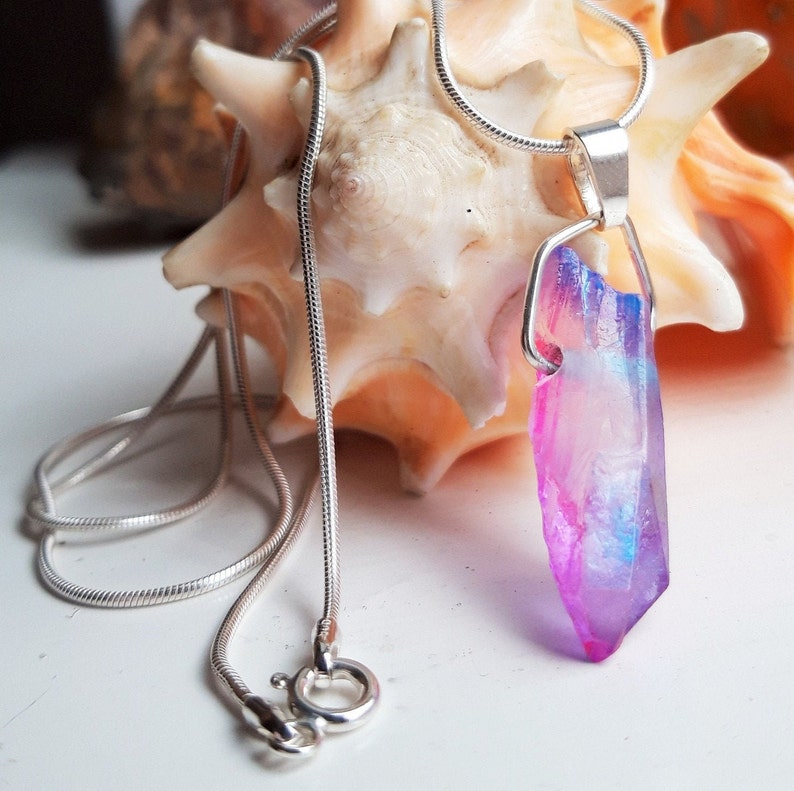 May include: A silver necklace with a pendant featuring a vibrant, multi-coloured crystal. The crystal displays shades of pink, purple, and blue. The necklace is displayed against a seashell background.