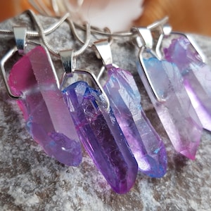 May include: Close-up of five crystal pendant necklaces. Each pendant features a raw, elongated crystal in shades of pink, purple, and blue, set in a silver-tone metal frame. The necklaces are displayed on a textured grey surface.